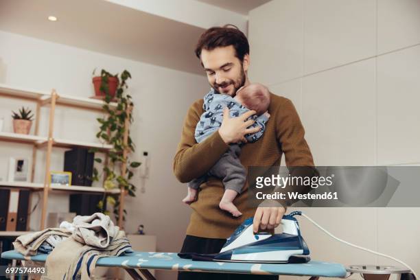 father ironing and holding his baby at home - multitasking stock-fotos und bilder