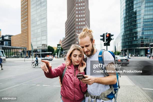 a young backpacking couple standing on corner of busy city street - viaje barato fotografías e imágenes de stock