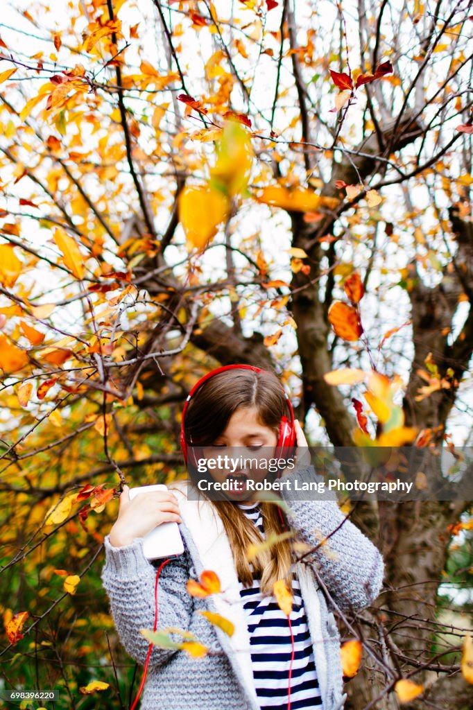 Young girl listening to music through headphones while outside under a colourful pretty tree during the fall