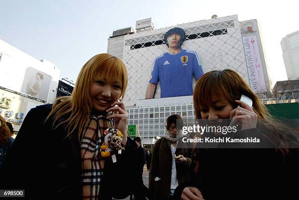 Two women speak on cellular telephones in front of a huge balloon depicting Japanese soccer player, Shunsuke Nakamura, wearing a team shirt for this...