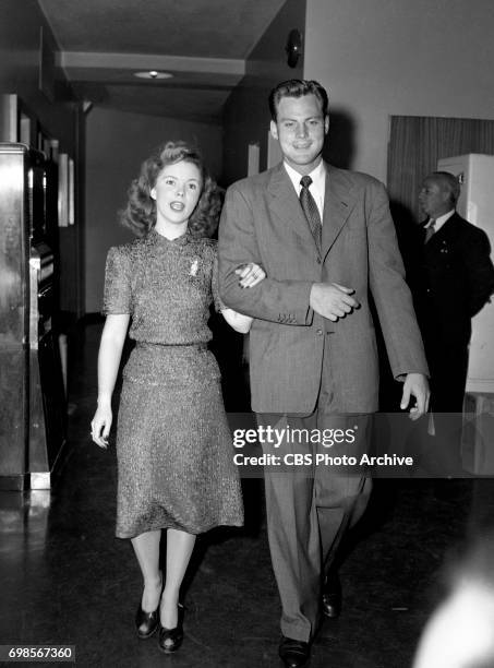 Mrs. And Mr. John Agar saunter through the corridor of Columbia Square to the broadcast studio. In background, at right, is George Temple, Shirleys...