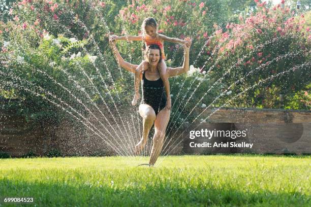 Mixed Race grandmother carrying granddaughter on shoulders in sprinkler