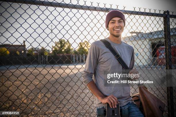 hispanic man leaning on chain-link fence with bag and skateboard - caldwell idaho stock pictures, royalty-free photos & images