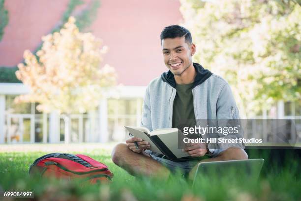 portrait of smiling hispanic man sitting in grass reading book - コールドウェル市 ストックフォトと画像
