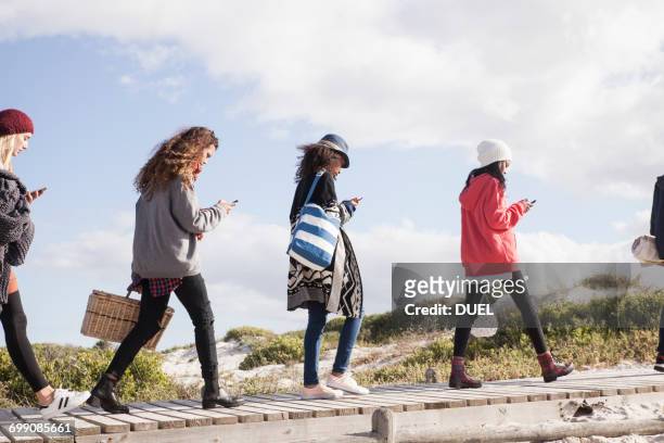 row of young adult friends strolling along beach boardwalk reading smartphones, western cape, south africa - obsession photos et images de collection