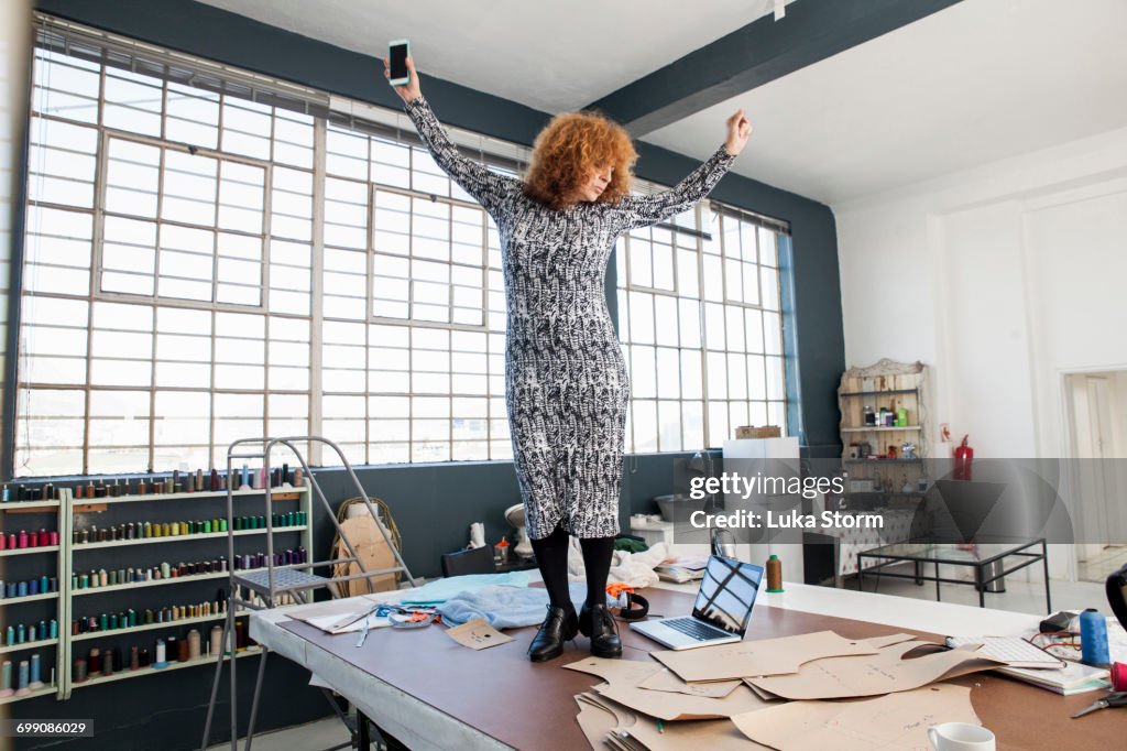 Mature female fashion designer dancing to smartphone music on workshop table