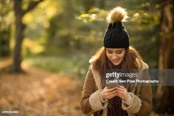 young woman in forest, looking at smartphone, smiling - bobble hat stock pictures, royalty-free photos & images