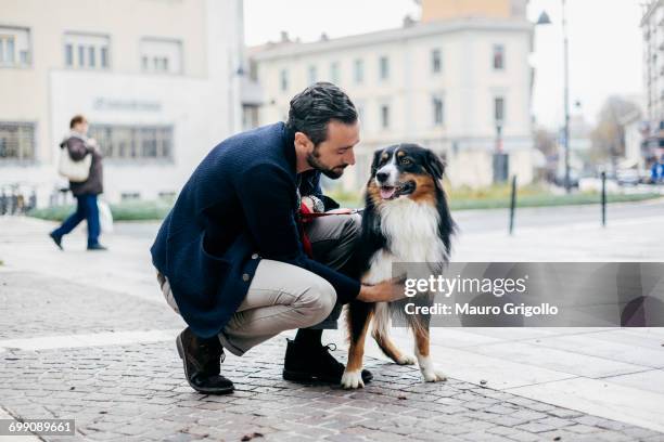 mid adult man crouching to pet dog in cobbled city square - travel loyalty stock pictures, royalty-free photos & images