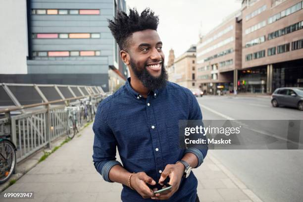 happy man with mobile phone while walking on sidewalk while looking away in city - coolpad stockfoto's en -beelden