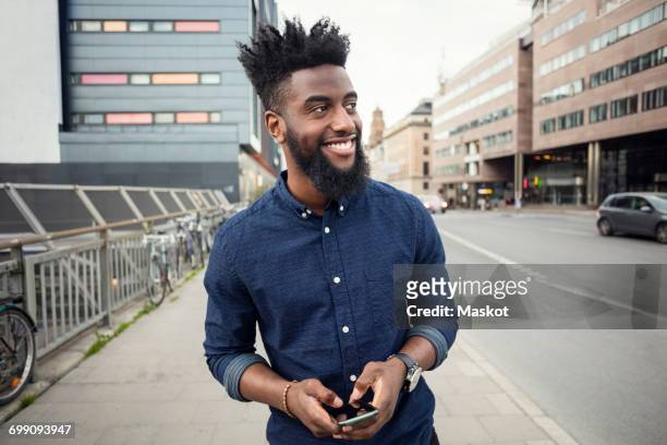 happy man with mobile phone while walking on sidewalk while looking away in city - authentic style stock pictures, royalty-free photos & images