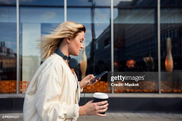 side view of woman using mobile phone while holding disposable cup against building - cup day one fotografías e imágenes de stock