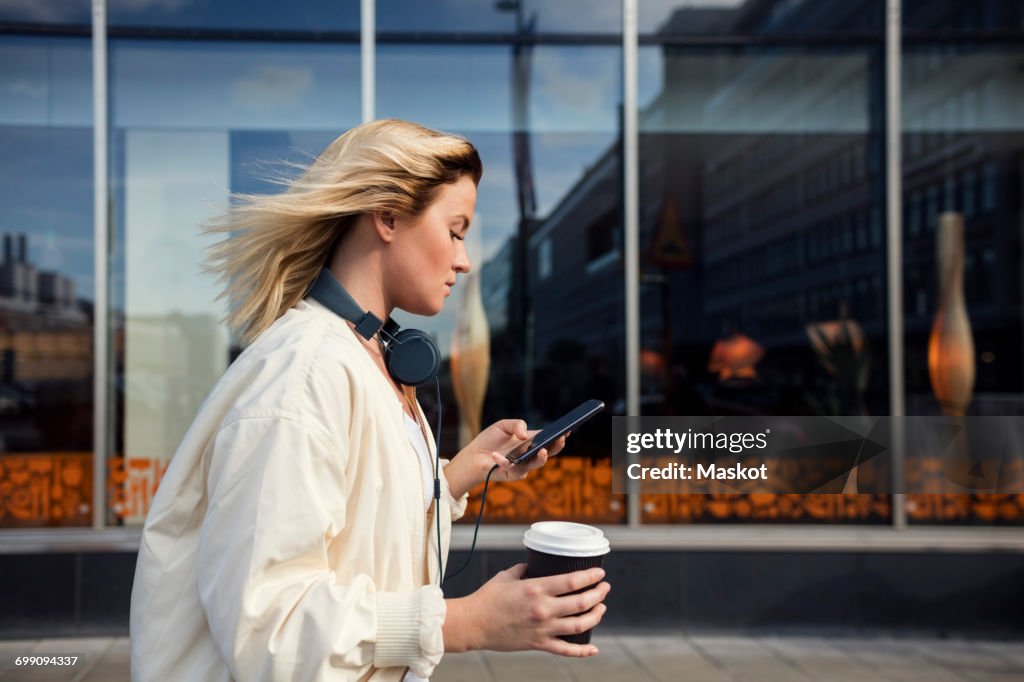 Side view of woman using mobile phone while holding disposable cup against building