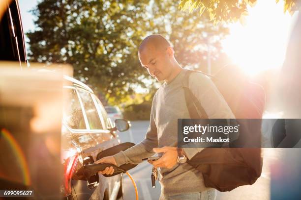 man charging car while using mobile phone at electric station on sunny day - vehículo de combustible alternativo fotografías e imágenes de stock