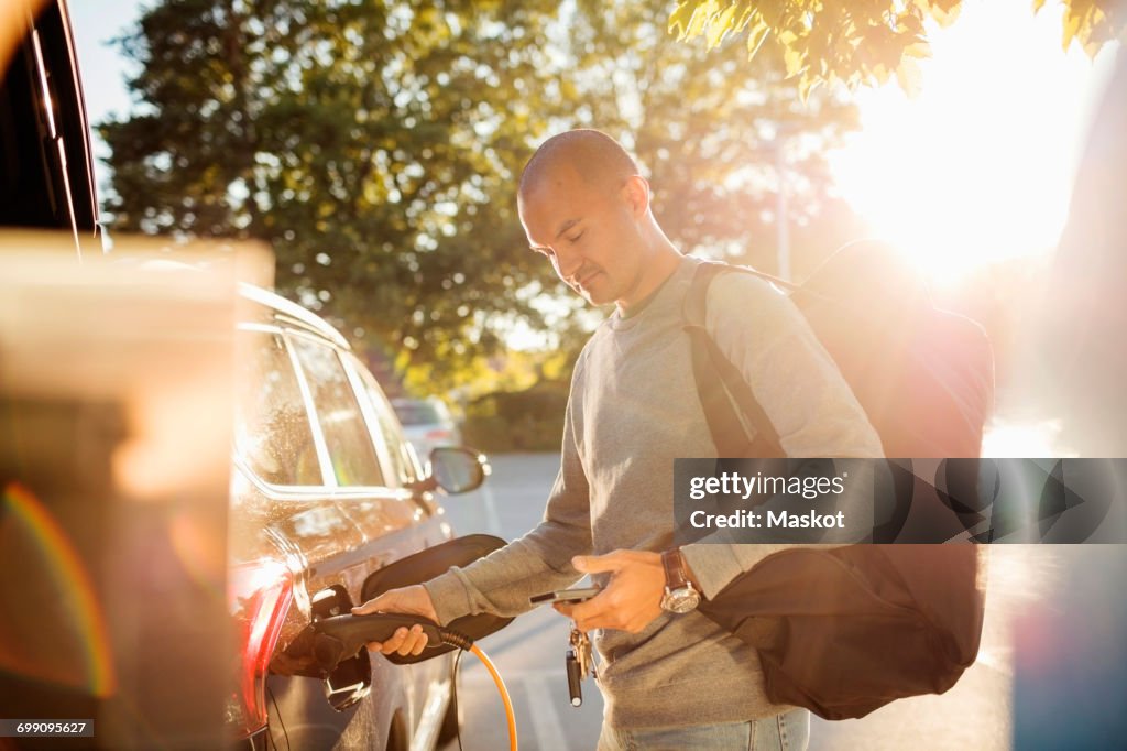 Man charging car while using mobile phone at electric station on sunny day