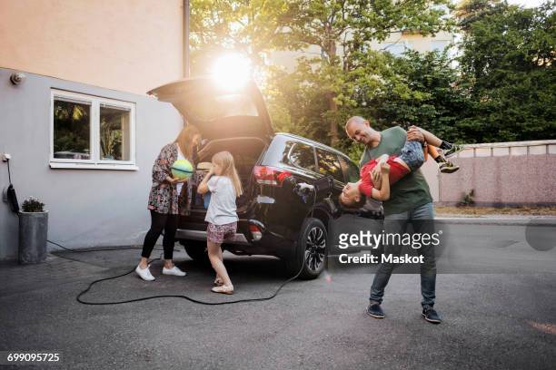playful father and son with woman and girl standing by car in back yard - father standing stockfoto's en -beelden