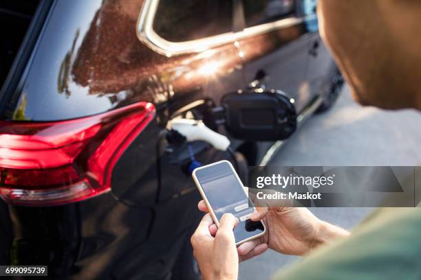 man using mobile phone while standing by charging electric car - car mobile stockfoto's en -beelden