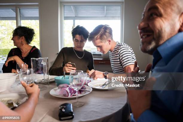 friends using phone while family talking at dining table in dinner party - immigrants refugees welcome fotografías e imágenes de stock