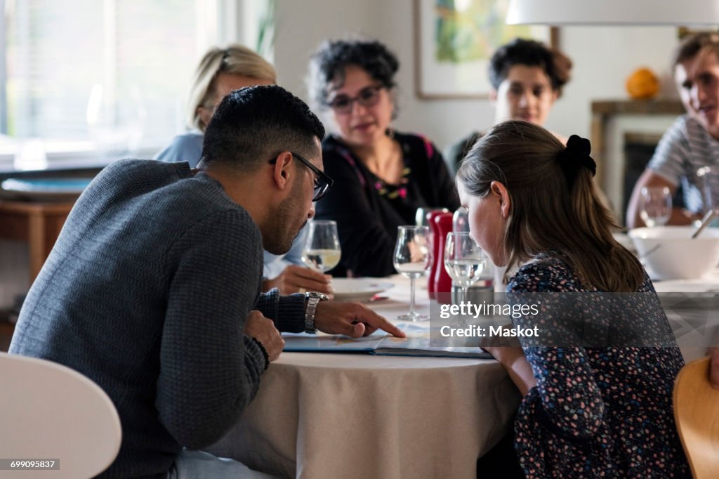 Man reading magazine to girl while sitting with friends and family at dinner party