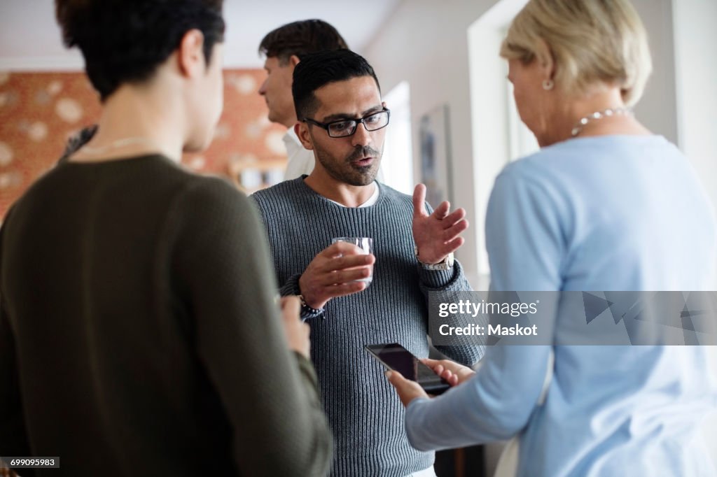 Man with glass gesturing while talking to woman holding phone at social gathering