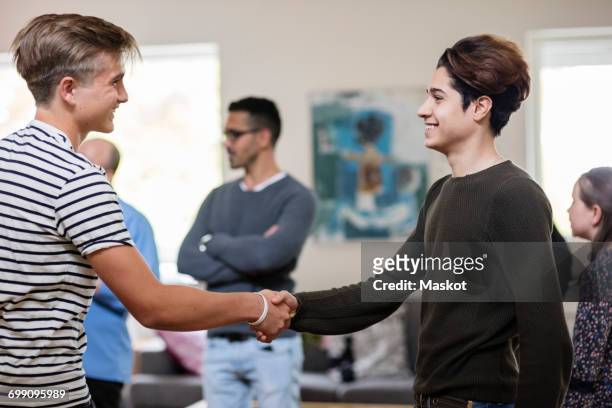 happy multi-ethnic friends shaking hands while standing with family in living room - reünie sociaal stockfoto's en -beelden