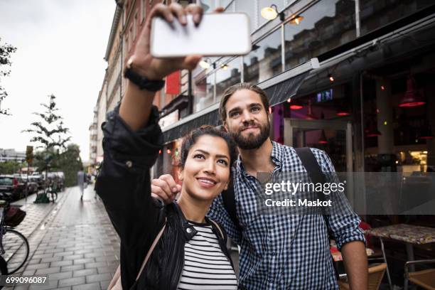 couple taking selfie through smart phone while standing by sidewalk cafe in city - pavement cafe - fotografias e filmes do acervo