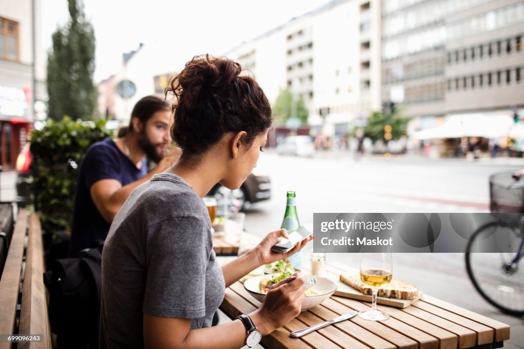 Woman having food while using mobile phone at sidewalk cafe in city