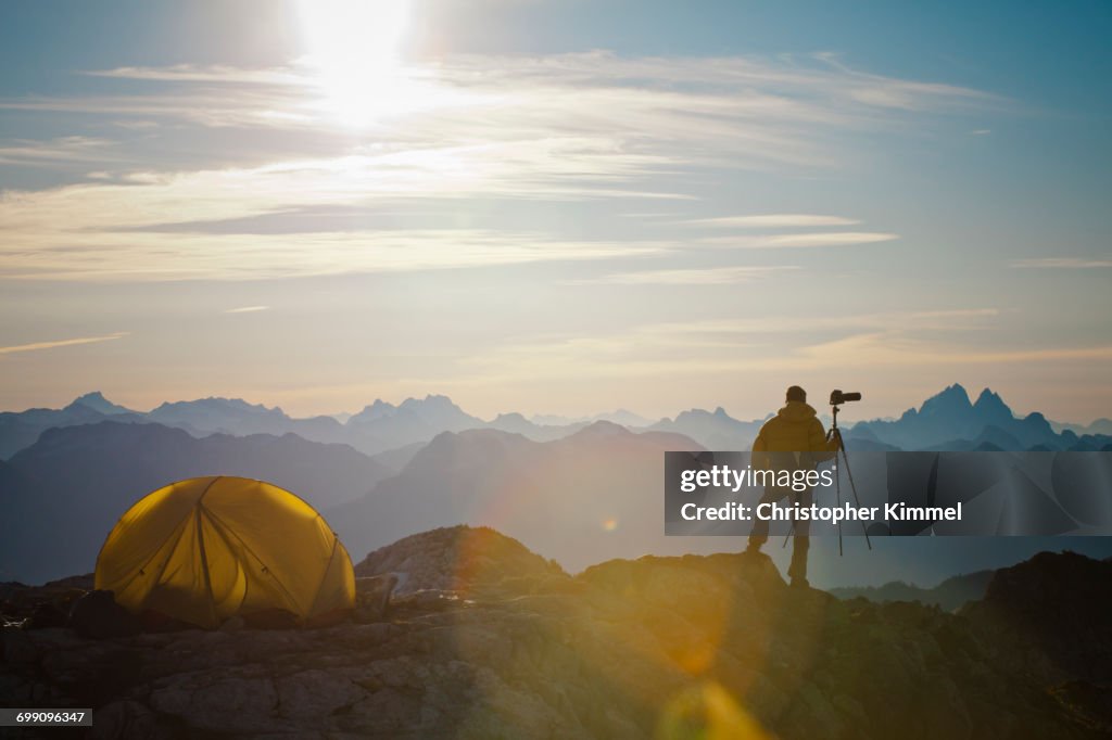 A photographer enjoys the view of the sunrise from his campsite on a rocky mountain ridge.