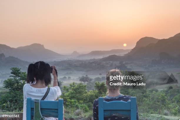 two girls sitting on wooden chairs watching the sunrise above vinales national park from los acuaticos, vinales, cuba - viñales cuba fotografías e imágenes de stock