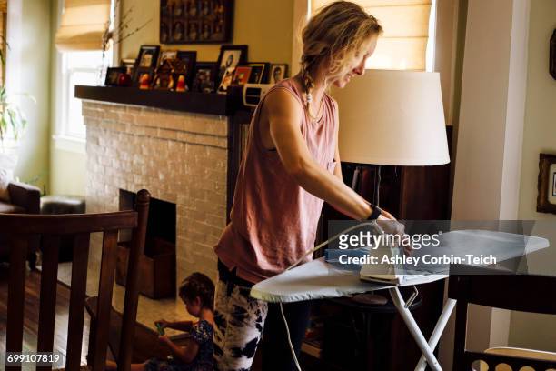 woman ironing whilst toddler daughter playing on floor - bügelbrett stock-fotos und bilder