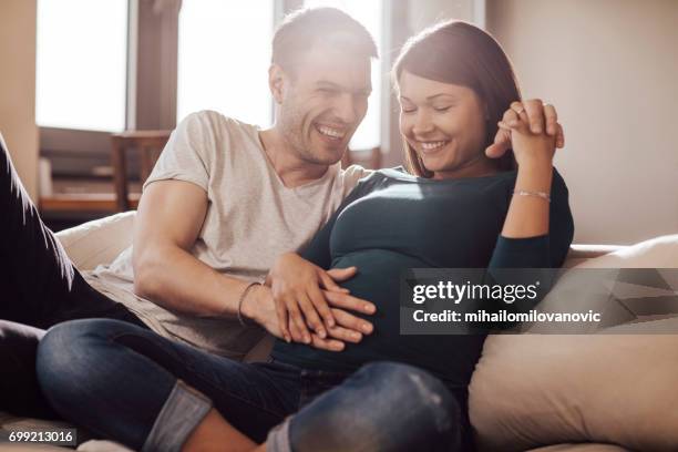 husband touching wife's stomach - young couple on couch stock pictures, royalty-free photos & images