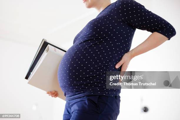 Berlin, Germany Posed scene: A pregnant woman is holding her aching back while she is holding file folders in the office on June 21, 2017 in Berlin,...