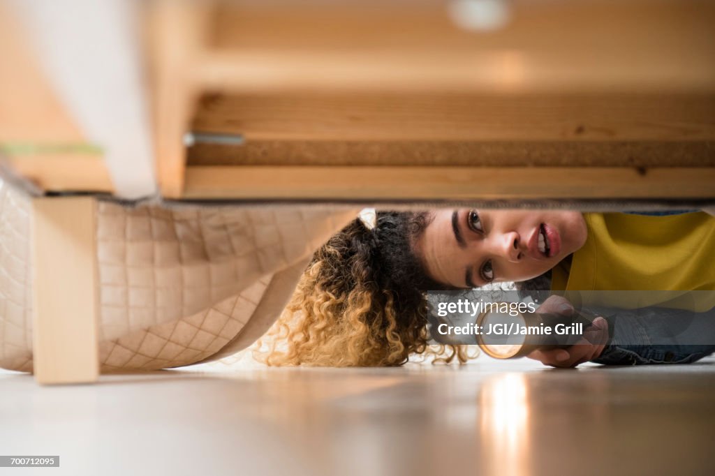 Mixed Race woman looking under bed with flashlight