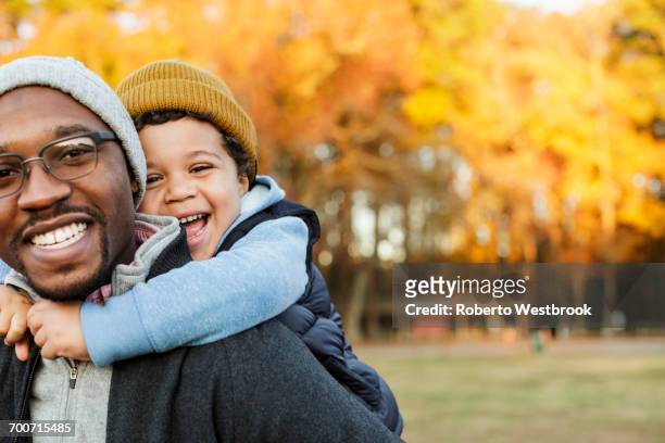 father carrying son piggyback in park - guancia a guancia foto e immagini stock