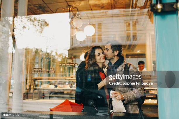 caucasian man kissing woman on cheek behind bakery window - bakery window stock pictures, royalty-free photos & images