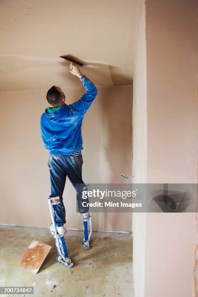 a plasterer wearing stilts smoothing fresh plaster high up on the walls of a house under construction. - plasterer stock pictures, royalty-free photos & images