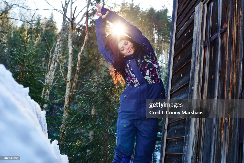 Woman in winter clothes practicing half moon yoga pose in snow by log cabin, Austria