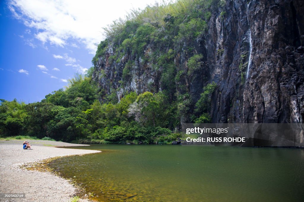 Couple sitting on tropical lakeside with rock waterfall, Reunion Island