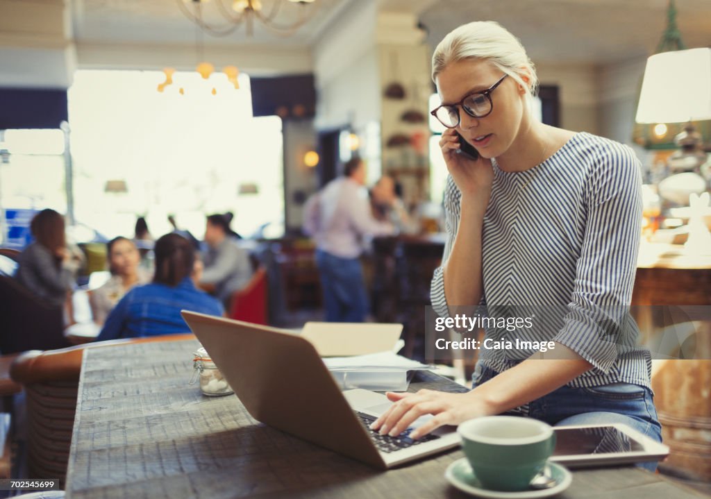 Creative businesswoman talking on cell phone and using laptop in cafe