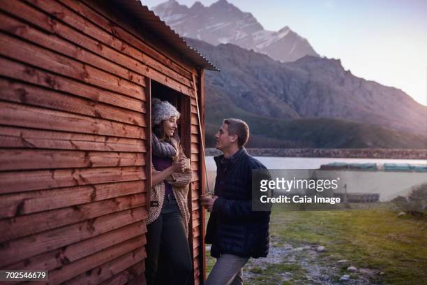 young couple drinking coffee at lakeside cabin doorway - vendeur stockfoto's en -beelden