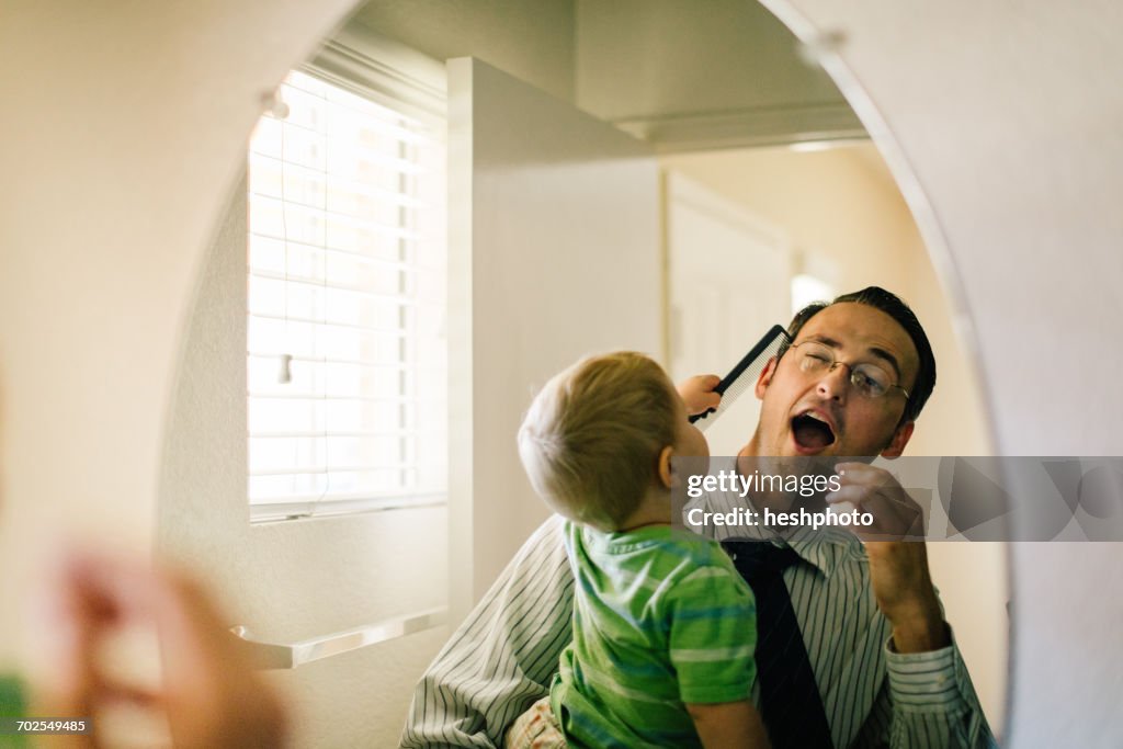 Father holding young son, son trying to comb fathers hair, reflected in mirror