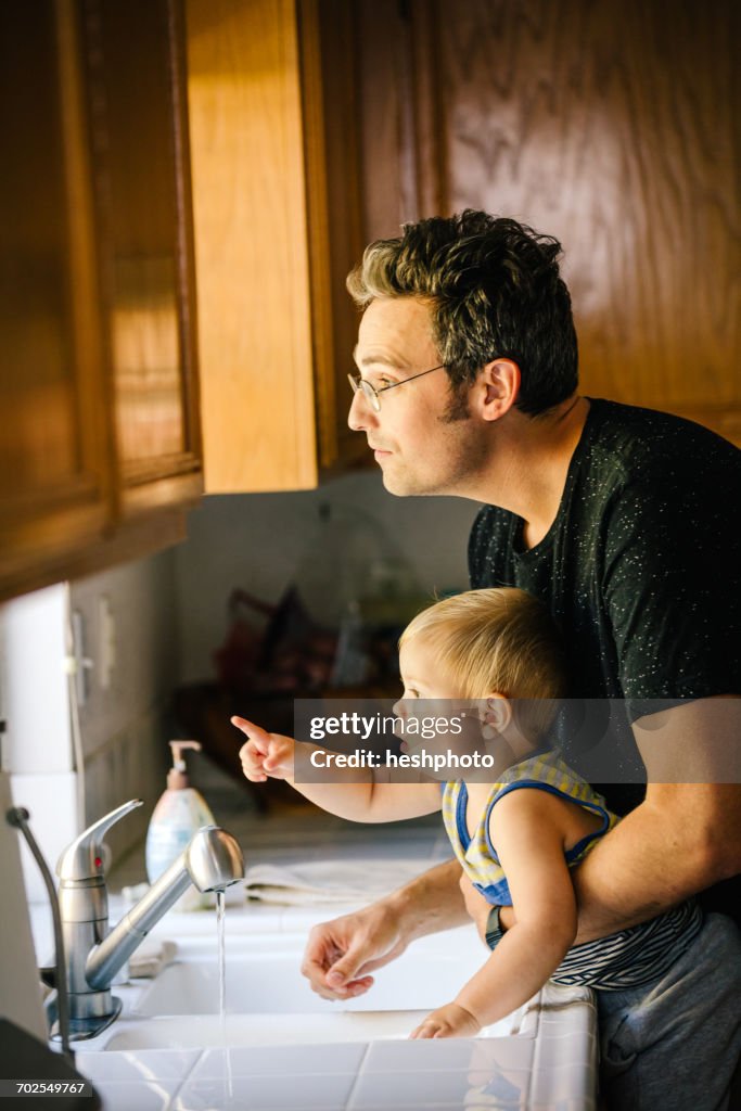 Father and young son washing hands at sink, looking out of window