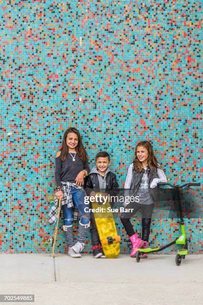 portrait of boy and two sisters with skateboards and scooter by multi-coloured urban wall - roller skates stock-fotos und bilder