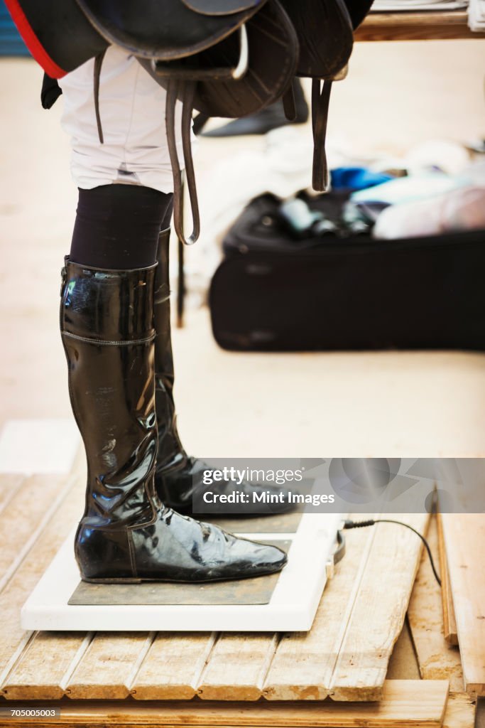 Rider wearing shiny black riding boots at the weigh in on weighing scale, holding a saddle, before or after a race.