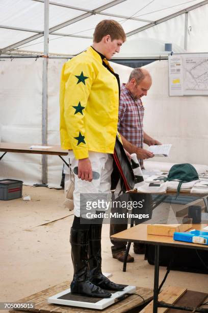 jockey in a yellow shirt standing on weighing scale, being weighed before a horse race. - jockey stock-fotos und bilder