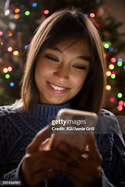 mixed race woman texting on cell phone near christmas tree - christmas smartphone stock-fotos und bilder