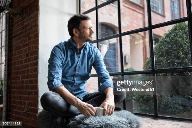 mature man sitting on window sill, relaxing with cup of coffee - apartamento tipo loft fotografías e imágenes de stock