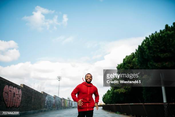 smiling young man wearing red hoodie running in the city - man running city stockfoto's en -beelden