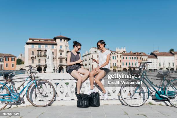 italy, padua, two young tourists sitting on railing looking at cell phones - city break friends stock pictures, royalty-free photos & images