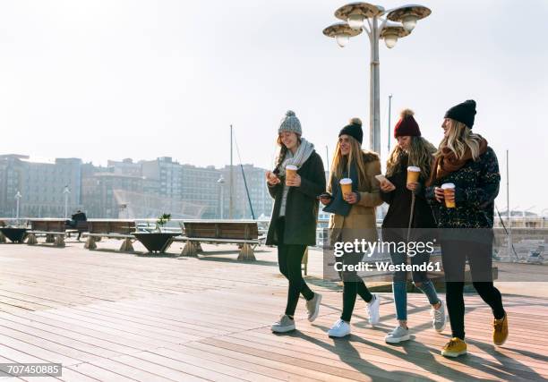 four friends with coffee to go and cell phones walking on promenade in winter - winter friends stock pictures, royalty-free photos & images