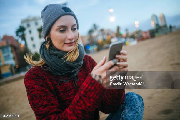 young woman sitting on the beach in winter using cell phone - winter scarf stock pictures, royalty-free photos & images